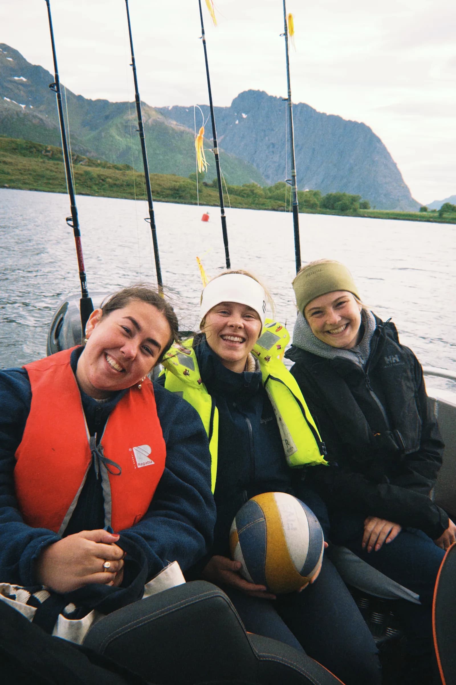 Tre elever smiler i båt med fiskestenger og fjell bak